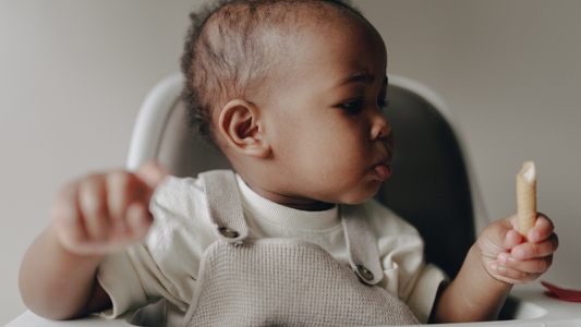 African- american baby sitting in a high chair with a piece of food in their hand.