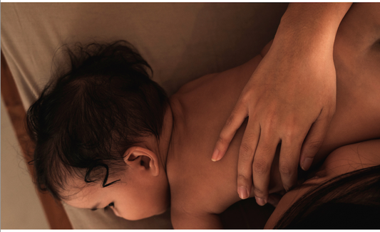baby laying down with a mother's hand over draped over its back in warm, ambient lighting
